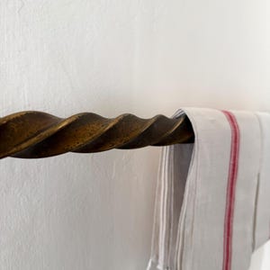 May include: A close-up of a bronze-coloured, twisted metal towel rail. A white towel with red stripes hangs from the rail. The rail is mounted on a white wall. The towel rail has a rustic, vintage aesthetic.