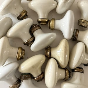 Puede incluir: Un conjunto de tiradores de armario antiguos de porcelana blanca con herrajes de latón. Los tiradores tienen un acabado liso y brillante y una forma redondeada clásica. Los herrajes de latón muestran signos de desgaste.