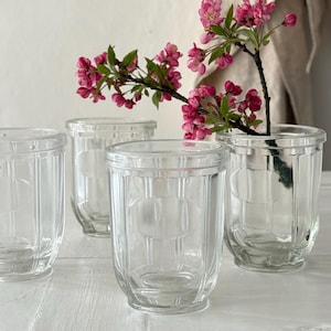 Puede incluir: Cuatro tarros de vidrio transparente con un diseño acanalado y un emblema ovalado. Un tarro contiene una rama con flores rosas. Los tarros están dispuestos sobre una superficie blanca, creando una exhibición simple y elegante.