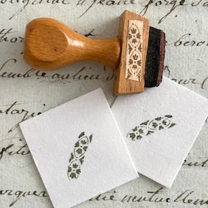 Op de afbeelding: Een rubberen stempel met een lichtbruin houten handvat en een rechthoekige basis met een decoratief bloemmotief. Er zijn twee kleine vierkante witte papiertjes aanwezig, elk met een groene inkt-afdruk van hetzelfde bloemmotief.