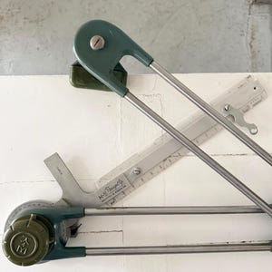 May include: A set of vintage drafting tools, including a green and silver parallel rule and a transparent ruler. The parallel rule has a circular dial with markings and the initials "M". The tools are set against a white background.