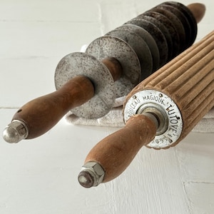 Può includere: Due utensili da cucina vintage: un tagliapasta con lame circolari e un mattarello scanalato. Entrambi hanno manici in legno e dettagli in metallo. Il mattarello presenta la scritta "ROULEAU MAGIQUE TUTOYE" all'estremità.