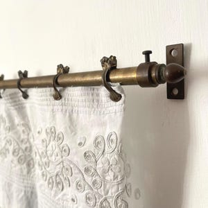Peut inclure: Une tringle à rideau en laiton vintage avec des embouts et des anneaux décoratifs, soutenant un rideau blanc brodé. La tringle est fixée sur un mur blanc, présentant un design de traitement de fenêtre classique.