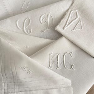 May include: Several white linen handkerchiefs, each featuring embroidered white monograms. The handkerchiefs are neatly folded, displaying the intricate stitching and diverse letter designs. The overall appearance is classic and refined, with a focus on detail.