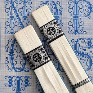 May include: Two spools of white DMC thread, labeled DMC 10 and DMC 16, are displayed against a blue and white patterned background. The thread spools have black and white labels with text. The background features a cross-stitch design.