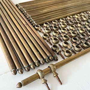 以下が含まれることがあります： アンティーク真鍮製の金具部品の詰め合わせ。ロッド、フィニアル、取り付けブラケットが含まれています。各部品は温かみのある経年変化が見られ、ヴィンテージまたは再生材を示唆しています。白い表面に配置され、各要素の詳細が際立っています。