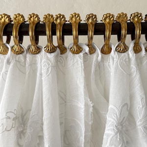 May include: Close-up of a curtain rod with ornate gold-colored curtain rings holding up white embroidered curtains. The curtains have a delicate floral pattern. The rod is dark brown.