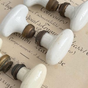 Può includere: Una collezione di pomelli per porta in porcellana bianca con componenti in ottone. Ogni pomello presenta un'impugnatura liscia di forma ovale fissata a una base metallica. La ferramenta mostra segni di usura con una patina scura e vissuta.