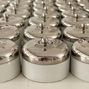 Op de afbeelding: Een verzameling vintage lichtschakelaars. Elke schakelaar heeft een glanzende, zilverkleurige metalen schakelaar en een witte keramische basis. De schakelaars zijn in rijen gerangschikt, wat wijst op een bulkverkoop of display.