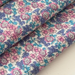 Op de afbeelding: Close-up van bloemenstof met een herhalend patroon van roze, blauwe en paarse bloemen op een crèmekleurige achtergrond. De stof is gevouwen en toont de textuur en details van het ontwerp. Ideaal voor naaien en knutselen.