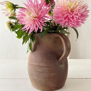 以下が含まれることがあります： 茶色の素朴な陶器のピッチャーに、ピンクと白のダリアの花束と緑の葉が入っています。花はとげのある花びらで、満開です。ピッチャーには取っ手があり、表面はざらざらしており、ヴィンテージ感があります。