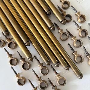 Op de afbeelding: Een verzameling vintage messing gordijnroeden en bijpassende hardware. De roeden zijn lang, cilindrisch en hebben puntige uiteinden. De hardware omvat oogringen met schroeven, allemaal in een warme, verouderde messing afwerking.