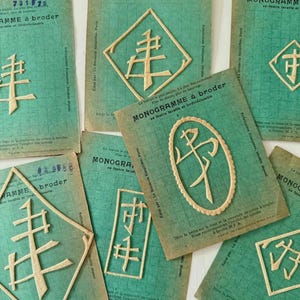 May include: A collection of vintage embroidery pattern cards. The cards are teal with a grid pattern and feature beige felt monograms in various geometric shapes. Text on the cards reads "MONOGRAMME à broder" and other French text.