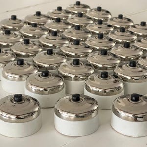 May include: A collection of vintage-style light switches. Each switch has a shiny, silver-colored dome and a black button, set on a white base. The switches are arranged in neat rows, suggesting a bulk purchase or display.