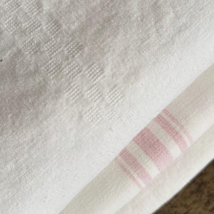 Peut inclure: Gros plan sur un tissu en lin blanc avec un motif carré texturé. Une section du tissu est pliée, révélant une section avec des rayures roses et blanches. Le tissu semble doux et légèrement froissé.