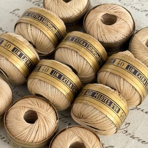 Könnte beinhalten: Eine Sammlung beigefarbener Leinengarnknäuel, jeweils umwickelt mit einem goldenen Papierband mit der Aufschrift LIN EXTRA 50 MÈTRES und 40. Das Garn ist zu kompakten, runden Formen gewickelt und weist eine gleichmäßige beige Farbe auf.