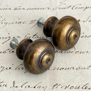 May include: Two round cabinet knobs with a dark, weathered brass finish. Each knob features a smooth, domed surface with a concentric circular design at the center and a short, threaded metal bolt extending from the back for installation.
