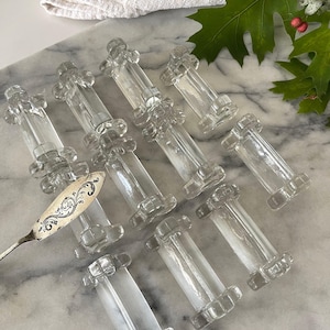 Puede incluir: Un conjunto de apoyacuchillos de vidrio transparente, cada uno con un cuerpo cilíndrico y extremos decorativos. Un cuchillo de mantequilla plateado con un diseño ornamentado también está presente. Los objetos están dispuestos sobre una superficie de mármol.