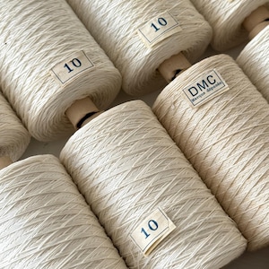 Op de afbeelding: Close-up van verschillende spoelen off-white garen. Elke spoel heeft een label met het nummer "10" erop gedrukt. Eén spoel heeft een label met de tekst "DMC Marque déposée". Het garen is strak gewikkeld.