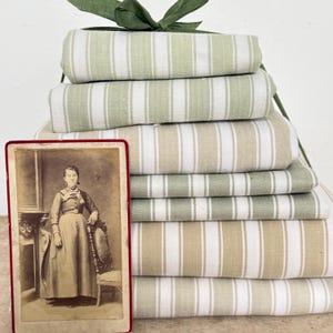 Può includere: Una pila di tessuti piegati con strisce verticali alternate in tonalità verde salvia, beige e bianco, legati insieme da un nastro verde. Una fotografia vintage color seppia di una persona in piedi accanto a una sedia è posta davanti.