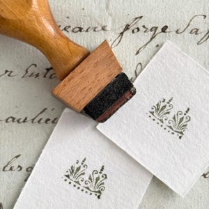 Op de afbeelding: Een houten stempel met een decoratief ontwerp wordt op wit papier gedrukt. De stempel heeft een bruine houten handgreep en een zwart inktkussen. Het gestempelde ontwerp is een groene bloemenkroon. De achtergrond bevat handgeschreven tekst.