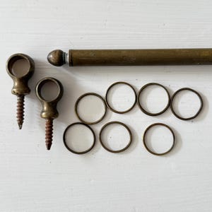 May include: An assortment of antique brass hardware components, including two eye-shaped screw-in mounts, a long rod with a decorative end, and several small rings. The items are arranged on a white surface.