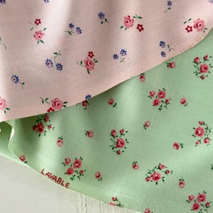 Pode incluir: Close-up de dois tecidos, um rosa claro e outro verde claro, ambos adornados com um delicado padrão floral. O tecido rosa apresenta pequenas rosas cor-de-rosa e flores azuis, enquanto o tecido verde tem rosas cor-de-rosa. A palavra "LAVABLE" está impressa no tecido verde.