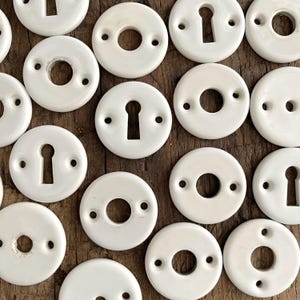 Può includere: Una collezione di bocchette per porte in porcellana bianca, alcune con fori per chiavi e altre con aperture circolari. Ogni bocchetta ha due piccoli fori per il montaggio. Oggetti disposti su una superficie di legno rustico.