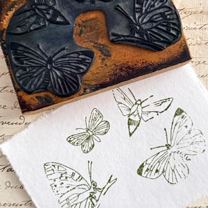 Op de afbeelding: Een houten stempel met een vlinderontwerp en een wit papier met groene vlinderopdrukken. De stempel heeft een donkerbruine basis met een zwart vlinderontwerp. Het papier heeft vijf verschillende vlinderontwerpen.