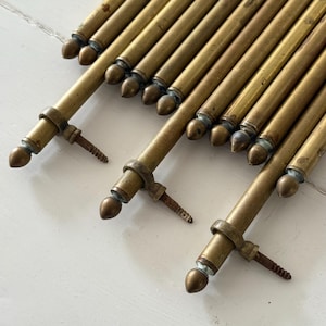 May include: Close-up of several antique brass rods with decorative finials and screw attachments. The rods are arranged diagonally, showcasing their aged patina and detailed craftsmanship. The warm, golden hue of the brass contrasts with the white surface.