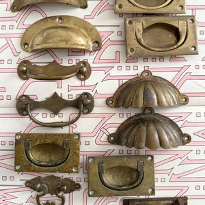 Op de afbeelding: Een verzameling antieke messing lade- en handgrepen in verschillende vormen en maten. De hardware omvat komtrekkers, valtrekkers en rechthoekige trekkers, allemaal met een verweerde, vintage uitstraling. De achtergrond is een roze en wit patroon.