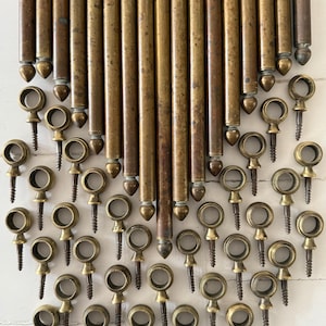 Peut inclure: Un assortiment de composants de quincaillerie en laiton vintage. L'image montre de longues tiges cylindriques avec des extrémités pointues, ainsi que de petites pièces avec des œillets circulaires et des vis filetées. Le laiton a une patine chaude et vieillie.