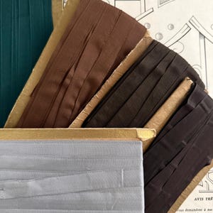Op de afbeelding: Een assortiment vintage lintspoelen in tinten van teal, bruin en grijs. De linten zijn netjes gerangschikt op kartonnen spoelen, waarbij hun texturen en kleuren worden getoond. De afbeelding legt de nadruk op de details van de stof en de verouderde uitstraling van de verpakking.