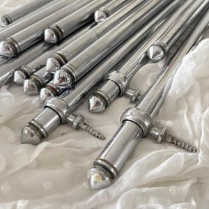 Puede incluir: Un conjunto de objetos metálicos plateados brillantes, con extremos puntiagudos y cuerpos cilíndricos. Los objetos tienen un diseño decorativo en el centro y un pequeño componente en forma de tornillo. Sobre tela blanca texturizada.