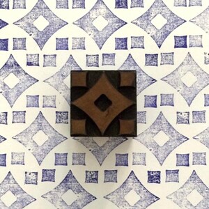 Può includere: Un timbro quadrato in legno con un disegno a rombo marrone al centro, circondato da dettagli neri. Il timbro è posto su una superficie bianca con un motivo a rombi blu ripetuto. Il motivo è composto da rombi e quadrati.