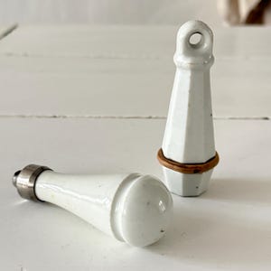 Könnte beinhalten: Zwei weiße Keramik-Schrankknöpfe mit Vintage-Ästhetik. Ein Knopf steht aufrecht und hat einen Ring oben und ein braunes Band. Der andere Knopf liegt, mit einem abgerundeten Ende und einer silbernen Metallbasis.