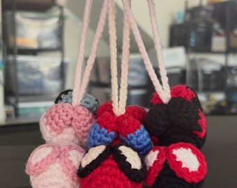 May include: Three crocheted yarn ornaments hanging from light pink braided cords. One ornament is light pink, one is red and blue with white eye shapes, and one is black and red with white eye shapes. Each piece features a round, textured design.