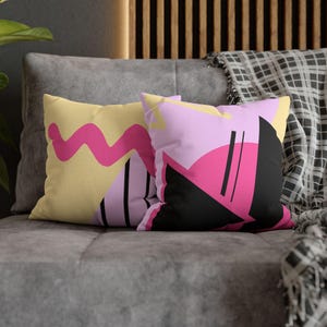Könnte beinhalten: Zwei dekorative Kissen mit abstrakten Designs. Ein Kissen hat einen gelben Hintergrund mit einem rosa Wellenmuster. Das andere Kissen hat einen rosa Hintergrund mit schwarzen, rosa und gelben geometrischen Formen. Die Kissen liegen auf einem grauen Sofa.