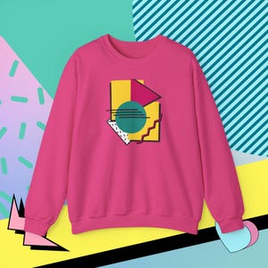 Op de afbeelding: Een magenta sweatshirt met een geometrisch ontwerp in geel, teal, roze en wit. Het ontwerp heeft een cirkel, driehoek en andere vormen omlijnd in zwart. De sweatshirt heeft lange mouwen en een ronde hals.