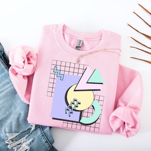 Könnte beinhalten: Ein hellrosa Sweatshirt mit einem geometrischen Design in Pastellfarben. Das Design umfasst ein Gitter, Dreiecke, einen Kreis und Zickzackformen. Eine zarte Halskette mit einem kleinen Anhänger liegt auf dem Sweatshirt.