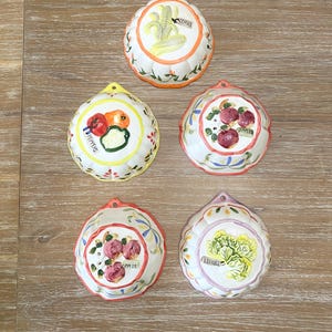 May include: Five ceramic wall-hanging molds with scalloped edges. Each features a hand-painted vegetable or fruit design with a corresponding label: CORNS, PEPPERS, APPLES, APPLES, and LETTUCE. The molds have white bases with colorful rims.