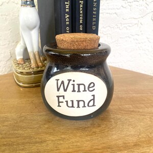 Pode incluir: Um pequeno pote de cerâmica redondo, de cor castanho-escuro, com acabamento brilhante e uma rolha de cortiça. A frente apresenta um rótulo oval branco com o texto "Wine Fund" impresso em letras castanho-escuras. O pote tem uma abertura larga.