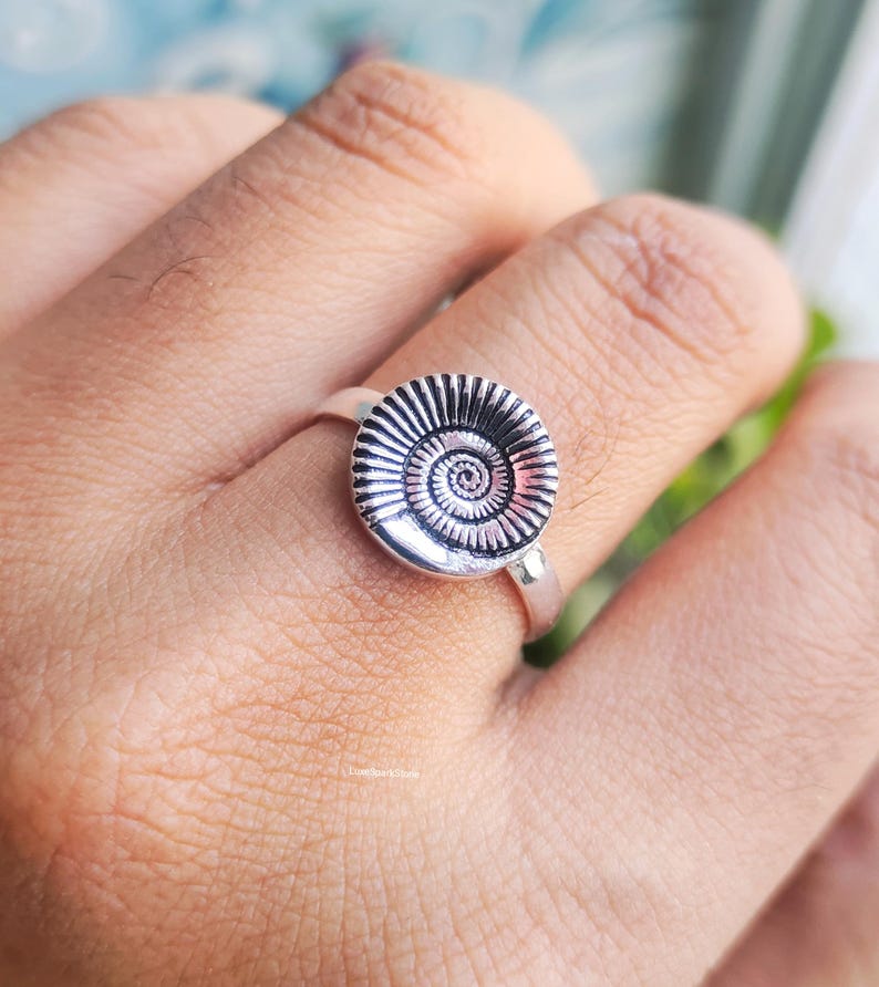 Pu&ograve; includere: Anello d'argento con un design circolare dettagliato che ricorda un fossile di ammonite. L'anello ha una fascia in argento lucido e il punto focale &egrave; il design a spirale e strutturato dell'ammonite. L'anello &egrave; indossato su un dito.