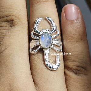 Könnte beinhalten: Ein silberner Skorpionring mit einem großen ovalen Mondstein-Edelstein. Das Design des Rings weist detaillierte Klauen, Körpersegmente und einen gebogenen Schwanz auf. Der Mondstein hat einen blau-weißen schillernden Glanz.