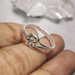 Könnte beinhalten: Ein silberfarbener Ring mit einem Knoten-Design. Der Ring hat ein Doppelband und einen detaillierten Knoten in der Mitte. Der Ring wird auf einer Hand gezeigt, mit einem unscharfen Hintergrund.