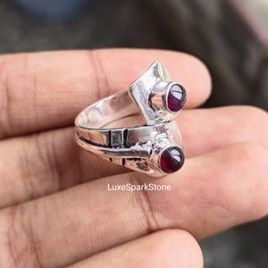 Könnte beinhalten: Ein silberner Ring mit zwei runden, tiefroten Edelsteinen. Der Ring hat ein einzigartiges Design mit geometrischen Formen und dem Text "LuxeSparkStone". Die Edelsteine sind in einer Zargenfassung gefasst.