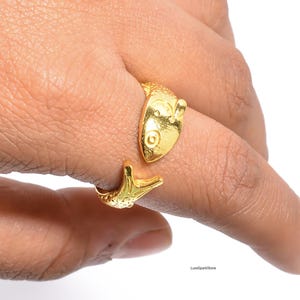 Könnte beinhalten: Ein goldfarbener Ring in Form eines Fisches mit detaillierten Schuppen und Flossen. Der Ring umschließt den Finger, wobei sich Kopf und Schwanz des Fisches treffen. Der Ring besteht aus einem glänzenden, reflektierenden Metall.