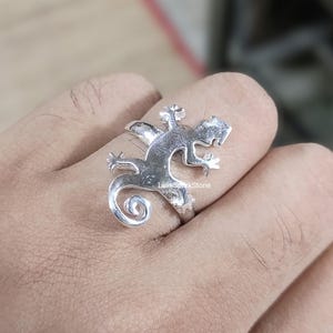 May include: A silver ring in the shape of a lizard, with a curled tail and detailed legs and head. The ring is worn on a finger, showcasing the intricate design and craftsmanship. The ring is a unique piece of jewelry.