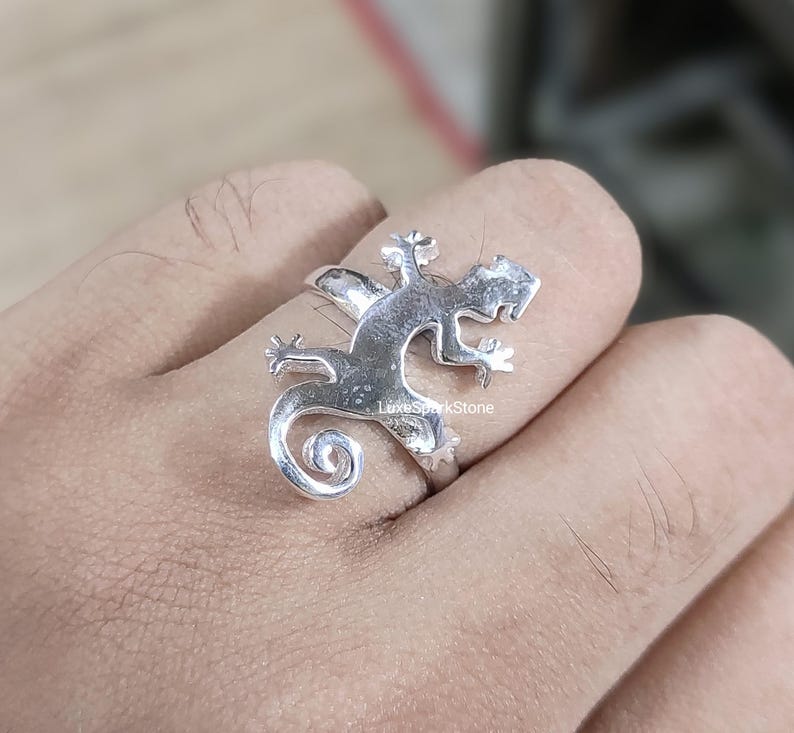 May include: A silver ring in the shape of a lizard, with a curled tail and detailed legs and head. The ring is worn on a finger, showcasing the intricate design and craftsmanship. The ring is a unique piece of jewelry.