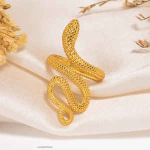 Könnte beinhalten: Ein goldfarbener Schlangenring mit strukturierter Oberfläche. Der Ring ist spiralförmig gewickelt, wobei der Schlangenkopf angehoben ist. Der Ring wird auf einer weißen, strukturierten Oberfläche präsentiert.
